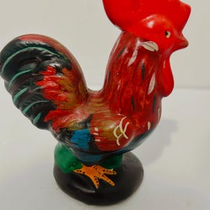 May include: A ceramic rooster figurine featuring a vibrant red comb and wattle, with a body painted in shades of red, black, blue, and green. The rooster stands on a solid black circular base, with yellow feet and white painted accents.