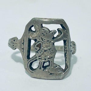 May include: A silver-toned metal ring featuring a rectangular face with a cut-out design of a cartoon mouse character. The mouse is depicted in a standing position with one arm raised. The ring band has a textured, decorative pattern on the sides.