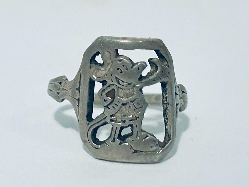 May include: A silver-toned metal ring featuring a rectangular face with a cut-out design of a cartoon mouse character. The mouse is depicted in a standing position with one arm raised. The ring band has a textured, decorative pattern on the sides.