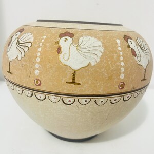 May include: A round ceramic bowl with a tan upper section featuring white rooster illustrations and vertical dot patterns. The lower section is a lighter cream color, separated by a dark brown line with a scalloped border design along the middle.