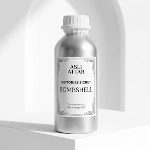 May include: A silver-colored bottle with a white cap and a label that reads "ASLI ATTAR VIKTORIES SICRET BOMBSHELL Concentrated Perfumery Oil." The bottle is cylindrical and sits on a white surface.