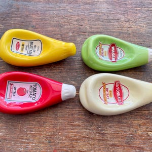 Könnte beinhalten: Vier Keramik-Dip-Schalen in Form von Gewürzflaschen: gelber Senf, grüner Wasabi, roter Ketchup und cremefarbene Mayonnaise. Jede Schale hat ein Etikett mit dem Namen des Gewürzes und eine weiße Spitze.