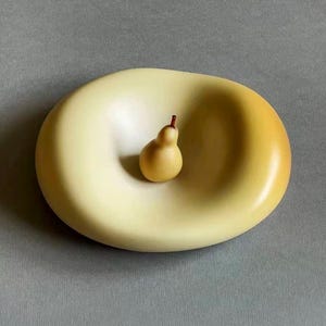 Pode incluir: Uma tigela decorativa amarela em forma de donut com um pequeno objeto em forma de pêra no centro. A tigela tem um acabamento liso e brilhante e um gradiente de cor do amarelo claro ao escuro. O objeto em forma de pêra tem um caule vermelho.