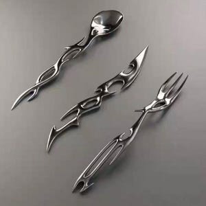 Puede incluir: Un juego de tres utensilios plateados: una cuchara, un cuchillo y un tenedor. Cada pieza presenta un diseño único y estilizado con bordes afilados y curvos y detalles calados. La cuchara tiene un cuenco redondeado, el cuchillo tiene una hoja puntiaguda y el tenedor tiene tres púas.