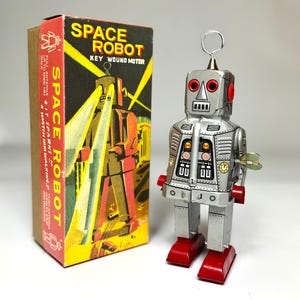 May include: A retro tin toy robot in silver with red details and a wind-up key, beside its original box. The box is labelled "SPACE ROBOT" with a vintage illustration. The robot has a wire antenna and red feet, evoking a nostalgic feel.