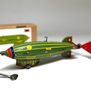 May include: A vintage green and yellow toy zeppelin with red and blue accents on the nose and a red propeller. The toy has a wind-up key and is displayed with its original box.