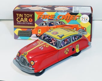 May include: Red and yellow wind-up tin toy car with a fire chief theme. The car has a yellow roof with a red and white siren on top. The car has a black and white F.D. logo on the side. The car is on a white background. The box for the toy car is in the background. The box is red, yellow, and blue with the words "Tin Toy Car Collections" and "Fire Chief" on it.