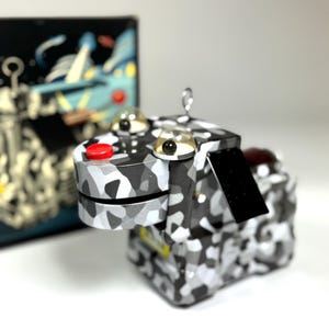 Könnte beinhalten: Ein Roboterspielzeug mit einem schwarz-weiß-grauen Tarnmuster. Das Spielzeug hat zwei klare, kuppelförmige Augen mit schwarzen Pupillen, einen roten Knopf und ein kleines schwarzes rechteckiges Stück. Im Hintergrund befindet sich eine Schachtel mit einer Weltraum-Illustration.