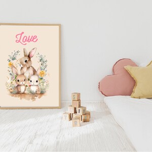 May include: A framed print with a light brown background featuring three bunnies with floral accents. The bunnies are brown and white with long ears. The word "Love" is written in pink at the top of the print.