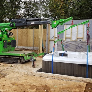 May include: A green tracked crane lifts a white hot tub into a wooden frame. The crane is operated by a person wearing a yellow safety vest and a white hard hat.