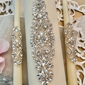 May include: Two ivory wedding candles with a decorative rhinestone and pearl design. The larger candle has a larger design with a more elaborate pattern.