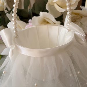May include: A white basket featuring a pleated satin interior and a handle adorned with white faux pearls. The exterior is wrapped in layers of sheer tulle fabric embellished with small white pearls, accented by satin bows on each side of the handle.