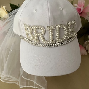 May include: White baseball cap with a veil attached. The cap has the word "BRIDE" spelled out in pearl-like beads with a rhinestone trim.