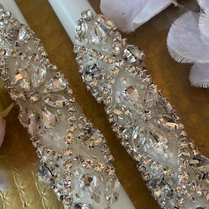 May include: Two white taper candles decorated with clear, faceted rhinestones arranged in a repeating floral and geometric pattern. The crystals are set in silver-toned metal settings, covering the middle section of each smooth, cylindrical candle.