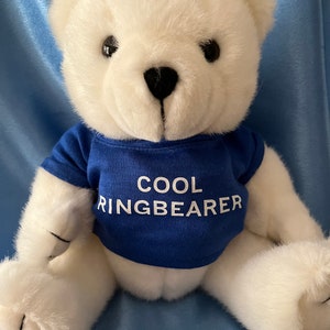 May include: White plush teddy bear wearing a blue t-shirt with the text "COOL RINGBEARER" in white.