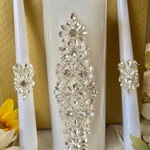May include: A set of three white wedding candles with decorative rhinestone embellishments. The center candle is larger and has a more elaborate design.
