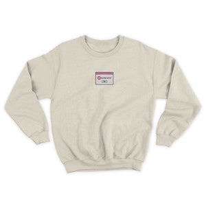 May include: A light beige sweatshirt with a small embroidered design of a pink and white computer error message. The message reads "System error".
