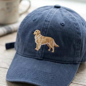 May include: A navy blue baseball cap featuring an embroidered golden retriever dog. The cap has a curved brim and an adjustable strap. The dog is depicted in a standing pose, with detailed fur and features.