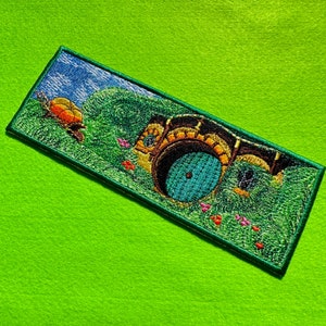 May include: Embroidered patch featuring a hobbit hole entrance with a round teal door, surrounded by green grass and a blue sky. A small orange and yellow creature is on the left. The patch has a green border.