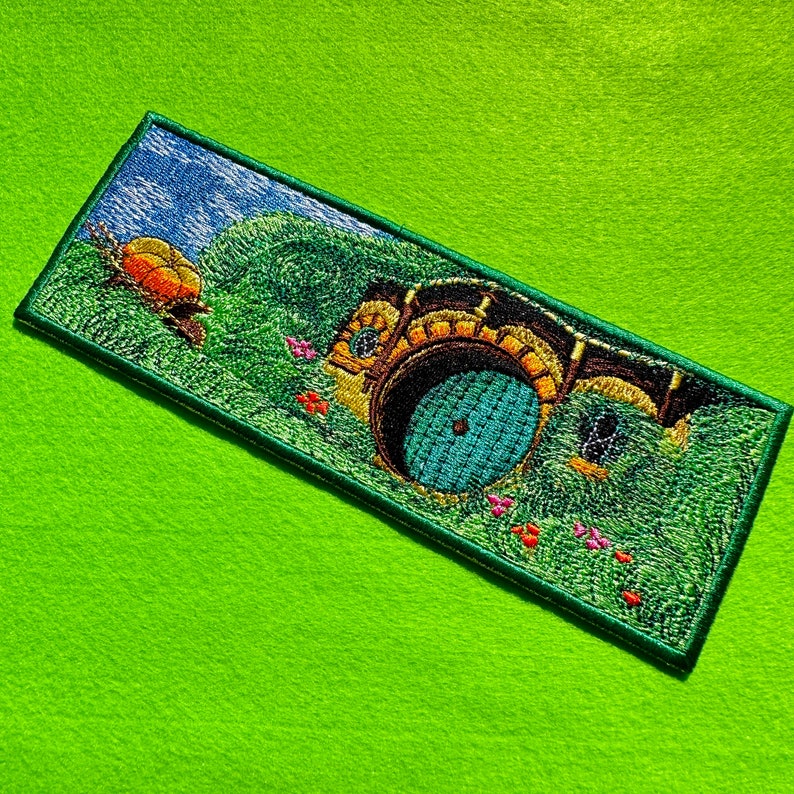 May include: Embroidered patch featuring a hobbit hole entrance with a round teal door, surrounded by green grass and a blue sky. A small orange and yellow creature is on the left. The patch has a green border.