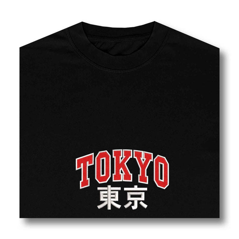 Puede incluir: Camiseta negra de cuello redondo con la palabra "TOKYO" en letras rojas con contorno blanco. Debajo, la traducci&oacute;n japonesa en blanco. La camiseta est&aacute; doblada.