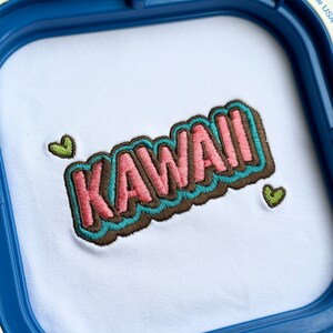 May include: Embroidered design of the word "KAWAII" in pink, blue, and brown with two green heart accents on a white fabric background.