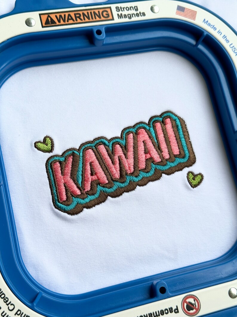 May include: Embroidered design of the word "KAWAII" in pink, blue, and brown with two green heart accents on a white fabric background.