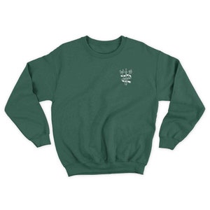 K&ouml;nnte beinhalten: Ein dunkelgr&uuml;ner Pullover mit einem wei&szlig;en gestickten Design auf der linken Brust. Das Design zeigt drei &Auml;xte mit einem Banner, auf dem "Good Things Take Time" steht.