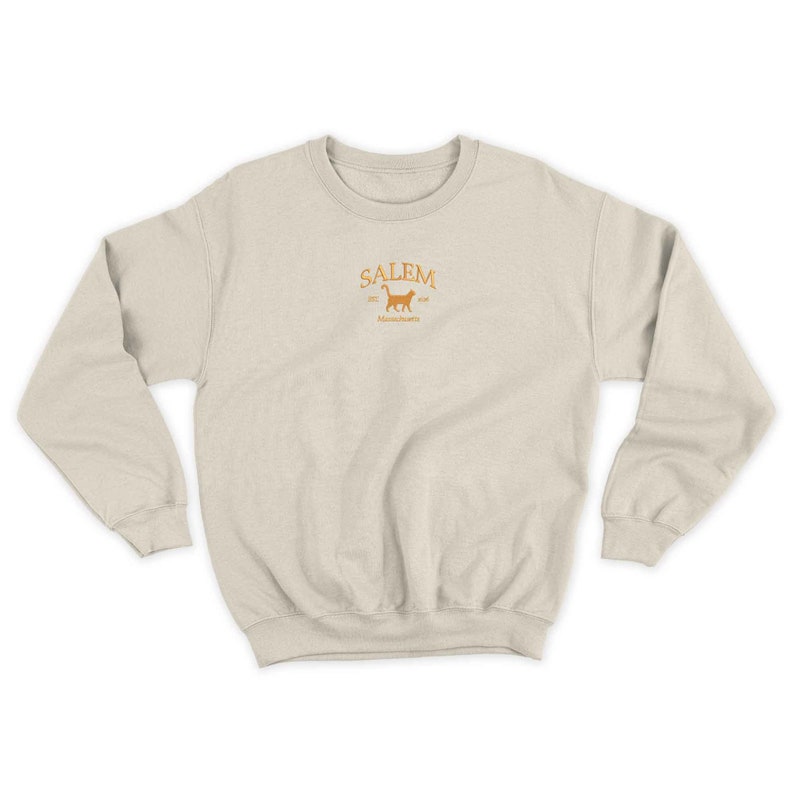 K&ouml;nnte beinhalten: Ein hellbeiger Pullover mit einem goldenen Stickmuster, das den Text "SALEM" mit einer Katzensilhouette und den Text "Massachusetts" darunter zeigt.