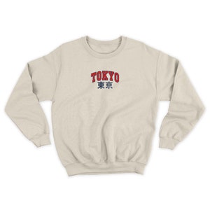Puede incluir: Sudadera color crema con la palabra "TOKYO" en rojo, contorneada en blanco, sobre caracteres japoneses grises. La sudadera tiene cuello redondo y mangas largas.