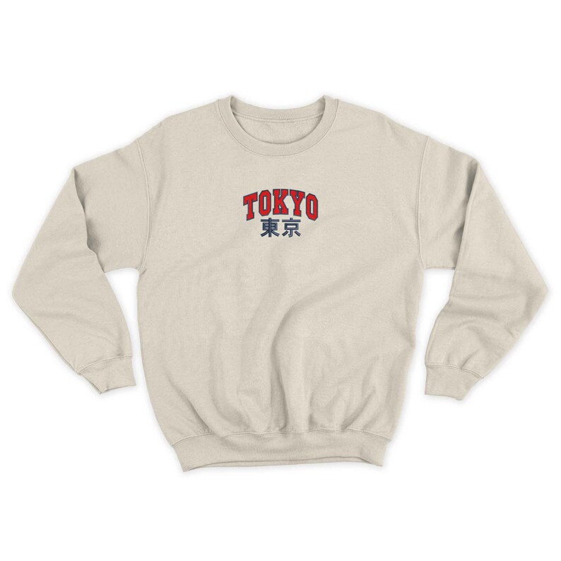 Puede incluir: Sudadera color crema con la palabra "TOKYO" en rojo, contorneada en blanco, sobre caracteres japoneses grises. La sudadera tiene cuello redondo y mangas largas.