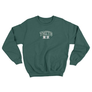 Puede incluir: Una sudadera verde oscuro con la palabra "TOKYO" en blanco, en letras de bloque sobre caracteres japoneses. La sudadera tiene cuello redondo y mangas largas.