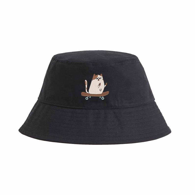 Op de afbeelding: Een zwarte bucket hat met een geborduurde afbeelding van een witte kat die skateboard.