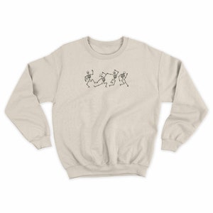 Pu&ograve; includere: Una felpa beige chiaro con una grafica di cinque scheletri che ballano. Gli scheletri sono in bianco e nero e sono in fila.