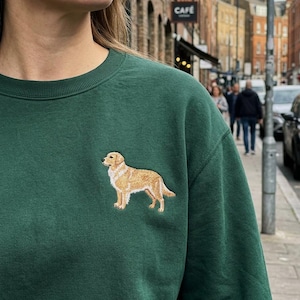 May include: A dark green sweatshirt featuring an embroidered golden retriever dog. The dog is depicted in a standing pose with detailed fur and features. The sweatshirt is a solid color, with a crew neck.