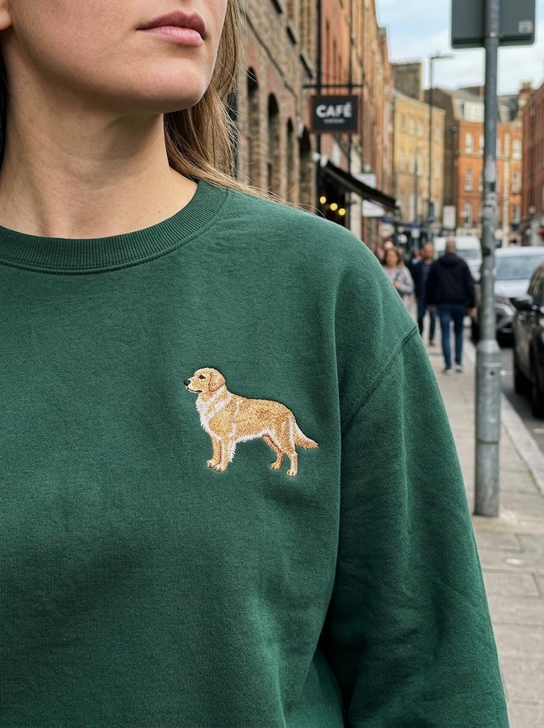May include: A dark green sweatshirt featuring an embroidered golden retriever dog. The dog is depicted in a standing pose with detailed fur and features. The sweatshirt is a solid color, with a crew neck.