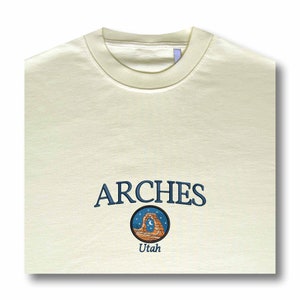 Pode incluir: Uma t-shirt de gola redonda cor de creme com o texto ARCHES bordado em azul, posicionado sobre um emblema circular que retrata um arco de rocha no deserto sob um c&eacute;u estrelado com uma lua crescente, com a palavra Utah bordada abaixo.