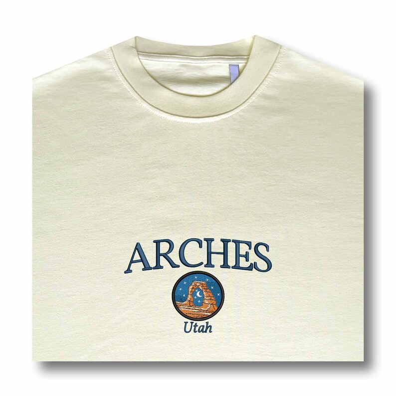 Pode incluir: Uma t-shirt de gola redonda cor de creme com o texto ARCHES bordado em azul, posicionado sobre um emblema circular que retrata um arco de rocha no deserto sob um c&eacute;u estrelado com uma lua crescente, com a palavra Utah bordada abaixo.