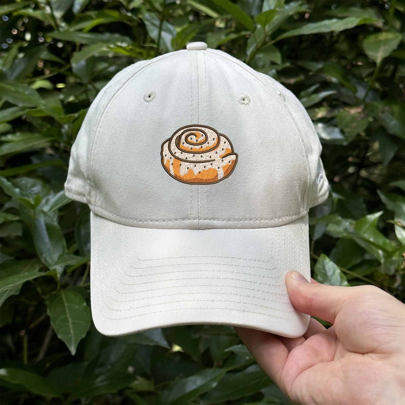 May include: A white baseball cap with a brown and white cinnamon roll design embroidered on the front.