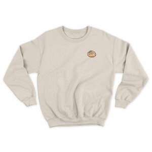 May include: A light beige sweatshirt with a small embroidered cinnamon roll on the left chest.