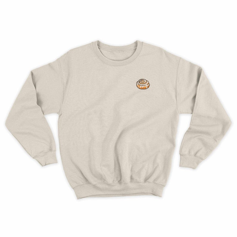 May include: A light beige sweatshirt with a small embroidered cinnamon roll on the left chest.
