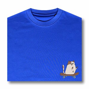 Op de afbeelding: Een koninklijk blauw t-shirt met een witte kat die op een skateboard rijdt. De kat heeft bruine vlekken en geeft een duim omhoog.