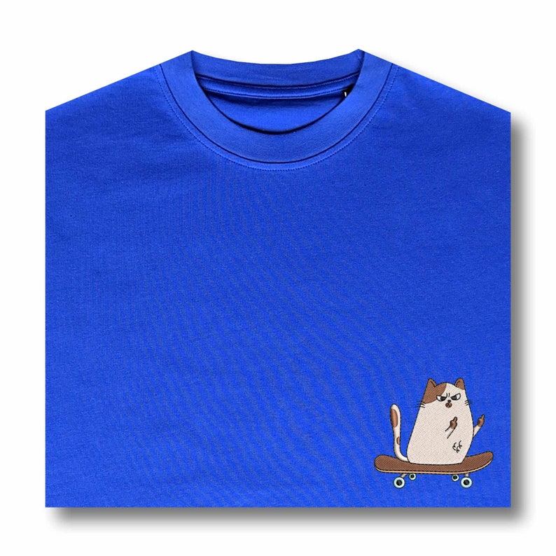 Op de afbeelding: Een koninklijk blauw t-shirt met een witte kat die op een skateboard rijdt. De kat heeft bruine vlekken en geeft een duim omhoog.