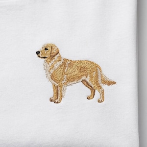 May include: Embroidered golden retriever dog design on a white t-shirt. The dog is depicted in a standing pose, with detailed stitching in shades of brown and tan. The shirt is made of a soft, white fabric.
