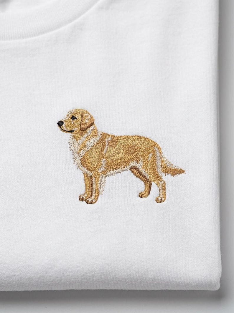 May include: Embroidered golden retriever dog design on a white t-shirt. The dog is depicted in a standing pose, with detailed stitching in shades of brown and tan. The shirt is made of a soft, white fabric.