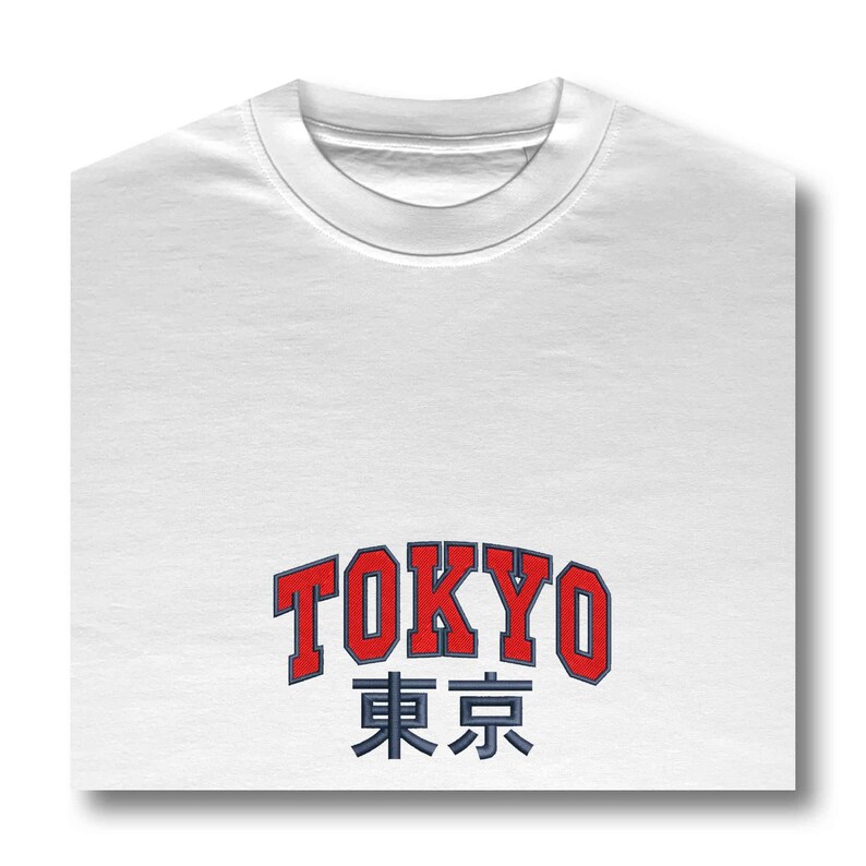 Puede incluir: Camiseta blanca con la palabra "TOKYO" en rojo, contorneada en azul, y caracteres japoneses debajo. La camiseta tiene cuello redondo y est&aacute; doblada cuidadosamente.