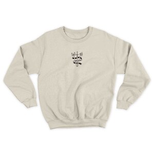 K&ouml;nnte beinhalten: Ein beiger Pullover mit einer schwarz-wei&szlig;en Grafik von drei gekreuzten Schwertern mit einem Banner, auf dem "Courage Under Fire" steht.