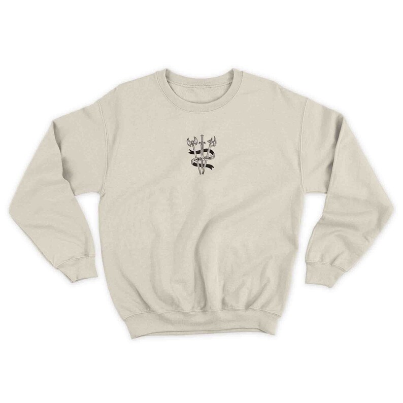 K&ouml;nnte beinhalten: Ein beiger Pullover mit einer schwarz-wei&szlig;en Grafik von drei gekreuzten Schwertern mit einem Banner, auf dem "Courage Under Fire" steht.