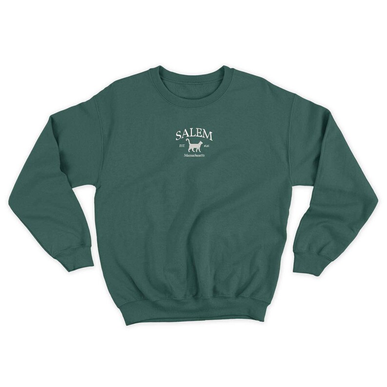 K&ouml;nnte beinhalten: Ein dunkelgr&uuml;ner Pullover mit einem wei&szlig;en gestickten Design auf der Vorderseite. Das Design zeigt das Wort "SALEM" mit einer Katzensilhouette und dem Text "est. 1626" und "Massachusetts".