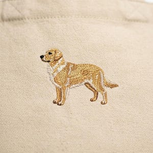 May include: Beige tote bag featuring an embroidered golden retriever dog. The dog is depicted in a standing pose with detailed stitching in shades of gold, brown, and white. The bag has a simple design with sturdy handles.